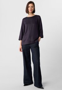 Top in satin viola scuro con scollatura a barca e maniche a tre quarti, abbinato a pantaloni in denim scuro a vita alta e a gamba larga e scarpe nere a punta.