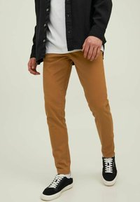 Tan slim-fit pants with a smooth texture, paired with a black shirt and black sneakers featuring white accents and laces.