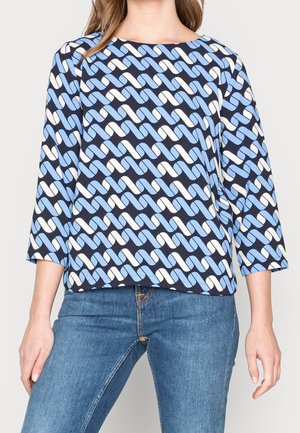 Femme portant un chemisier à motifs géométriques noirs, blancs et bleus avec des manches trois-quarts et un jean en denim bleu.