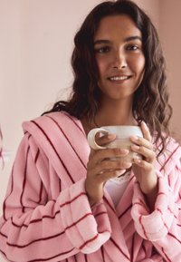 Pink striped bathrobe with plush texture; oversized sleeves. The person holds a textured, light-colored mug, standing against a pale background.