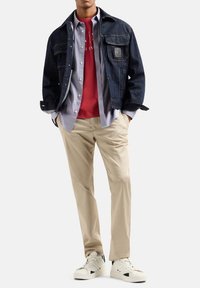 Giacca in denim scuro, camicia grigia chiara con bottoni, t-shirt rossa, pantaloni beige e sneaker bianche. Presenta tasche e accenti in contrasto.