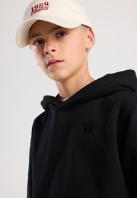 Felpa nera con tasca a marsupio, caratterizzata da un piccolo logo ricamato. Indossata con un cappellino crema che presenta testo rosso "1989 Classics."