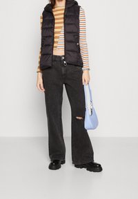 Zwarte puffer vest, gestreept lang mouwtop met meerkleurige strepen, zwarte wijde jeans met een knie-scheur, en een kleine lichtblauwe handtas.