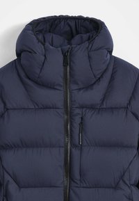 Veste matelassée marine avec col montant, motif cousu et fermeture éclair noire. Texture douce et isolation légère visible.