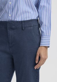 Kék farmer nadrág sima textúrával és karcsúsított szabással, világoskék és fehér csíkos inggel párosítva, gombos részletekkel.