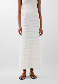 Longa gonna bianca in maglia con strisce orizzontali testurizzate e dettagli decorativi. Indossata con sandali neri con cinturini.