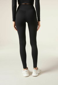 Svarta högmidjade leggings med en mjuk, elastisk textur, kombinerade med ljusa sneakers, visar en åtsittande design bakifrån.