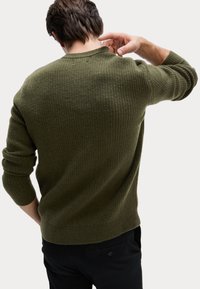 Olivgrön stickad tröja med en texturerad mönster, ribbade ärmslut och rundad hals; kombinerad med svarta byxor, vy från baksidan.