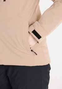 Beige jacka med texturerad yta, som har justerbara ärmslut med svart kardborre och en sidoficka med dragkedjedetalj.