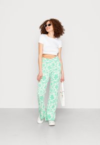 Calças verde menta com corte flare e padrão floral branco, combinadas com uma t-shirt cropped branca e ténis brancos, complementadas por uma bolsa branca.