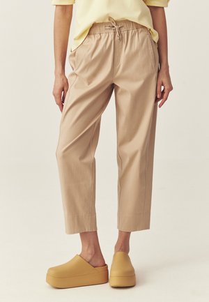 Osoba ubrana w beżowe spodnie ze ściągaczem i żółte buty na platformie wsuwane, stojąca na jednolitym jasnym tle.