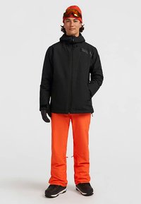 Veste imperméable noire avec fermeture éclair, pantalon imperméable orange, gants noirs et lunettes de ski à teinte orange. Convient pour temps froid.