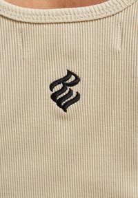 Beige ribbet stof med et broderet sort logo med bølgede linjer, placeret centralt. Udhullet er rundt og struktureret.