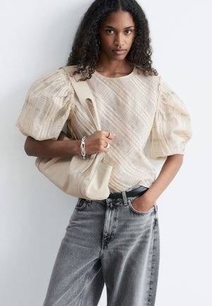 Žena s kovrčavom kosom u bež majici s naboranim rukavima i sivim trapericama, drži bež torbu na ramenu s jednom rukom u džepu.