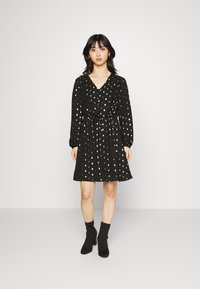 Vestido preto com mangas longas, decote em V, cintura ajustada e bolinhas prateadas. Textura suave, comprimento acima do joelho, combinado com botas pretas até o tornozelo.