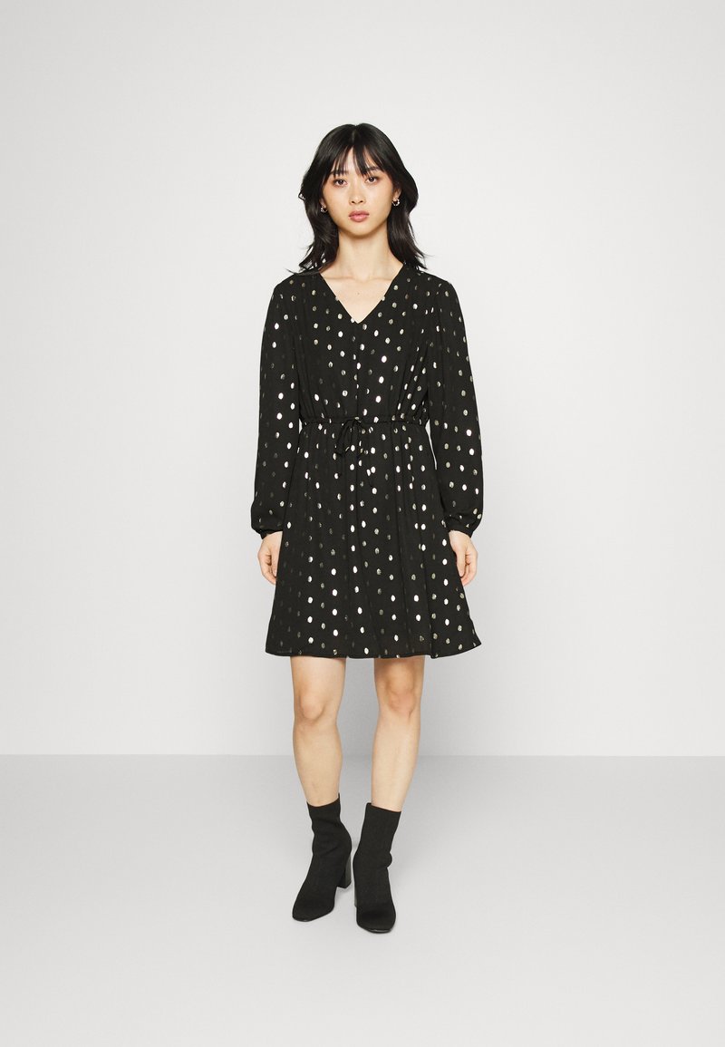 Vestido preto com mangas longas, decote em V, cintura ajustada e bolinhas prateadas. Textura suave, comprimento acima do joelho, combinado com botas pretas até o tornozelo.