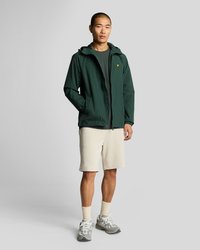 Žalia neperšlampama striukė su gaubtu, priekiu su užtrauktuku ir elastingais rankogaliais, derinama su kreminėmis šortais. Dėvimi pilki sportbačiai ir balti kojinai.