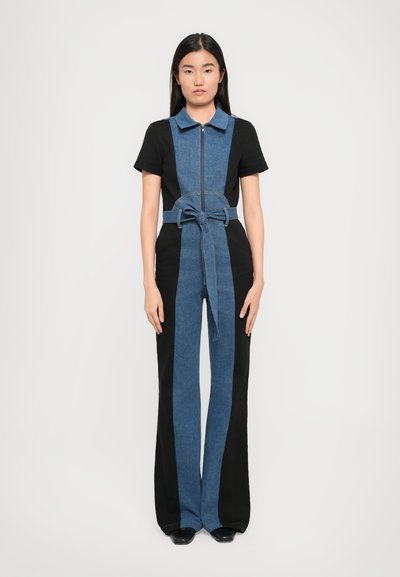 Femme debout portant une combinaison en denim noire et bleue à manches courtes, avec une fermeture éclair à l'avant, un col et une ceinture nouée à la taille, sur un fond blanc.