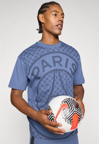 Mėlynas medvilninis marškinėlis su tekstūruotu grafinio rašto ir užrašu "PARIS". Asmuo laiko baltą ir juodą Nike futbolo kamuolį su raudonais akcentais.
