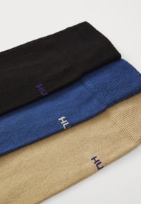 Drei Paare Socken flach ausgelegt: schwarz, blau und beige. Jede hat einen gerippten Bund und zeigt ein kleines Logo in kontrastierender Farbe.