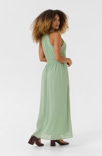 Maxi dress in light green, sleeveless with a gathered waist, flowing fabric, and a smooth texture; paired with dark brown chunky heels.