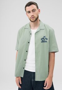 Light green short-sleeve shirt in a textured fabric with a button front. Features navy blue embroidered text "HERE FOR THE BEER."