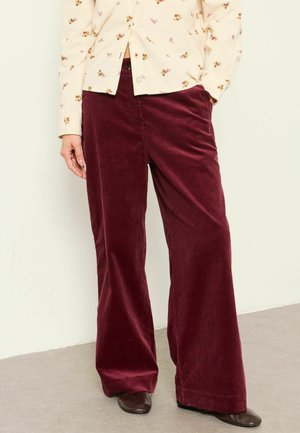 Person wearing wide-leg burgundy corduroy pants, cream floral long-sleeve shirt, and dark brown shoes standing on a light floor.