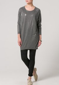 Šedý tunikový top se flitry, kulatým výstřihem a volným střihem, doplněný černými legínami s vzorováním a béžovými botami. Měkká textura látky.