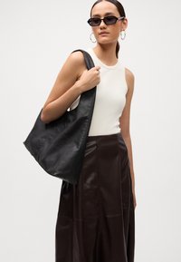 Geantă tote din piele neagră cu un design lejer, asortată cu un top alb mulat și o fustă din piele maro. Modelul poartă ochelari de soare închiși la culoare.