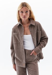 Bruine corduroy overhemd met kraag, voorzak en knoopsluiting, gecombineerd met bijpassende wijde broeken en een wit tanktop eronder.
