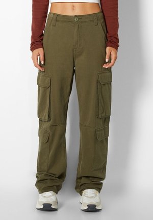 Vrouw draagt losse olijfgroene cargobroek met meerdere klepzakken, witte sneakers en een cropped roestkleurige longsleeve top.