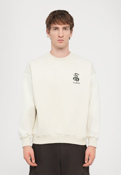 Sudadera color crema hecha de material suave, con un pequeño logo de serpiente negra y puños y dobladillo acanalados, combinada con pantalones oscuros.