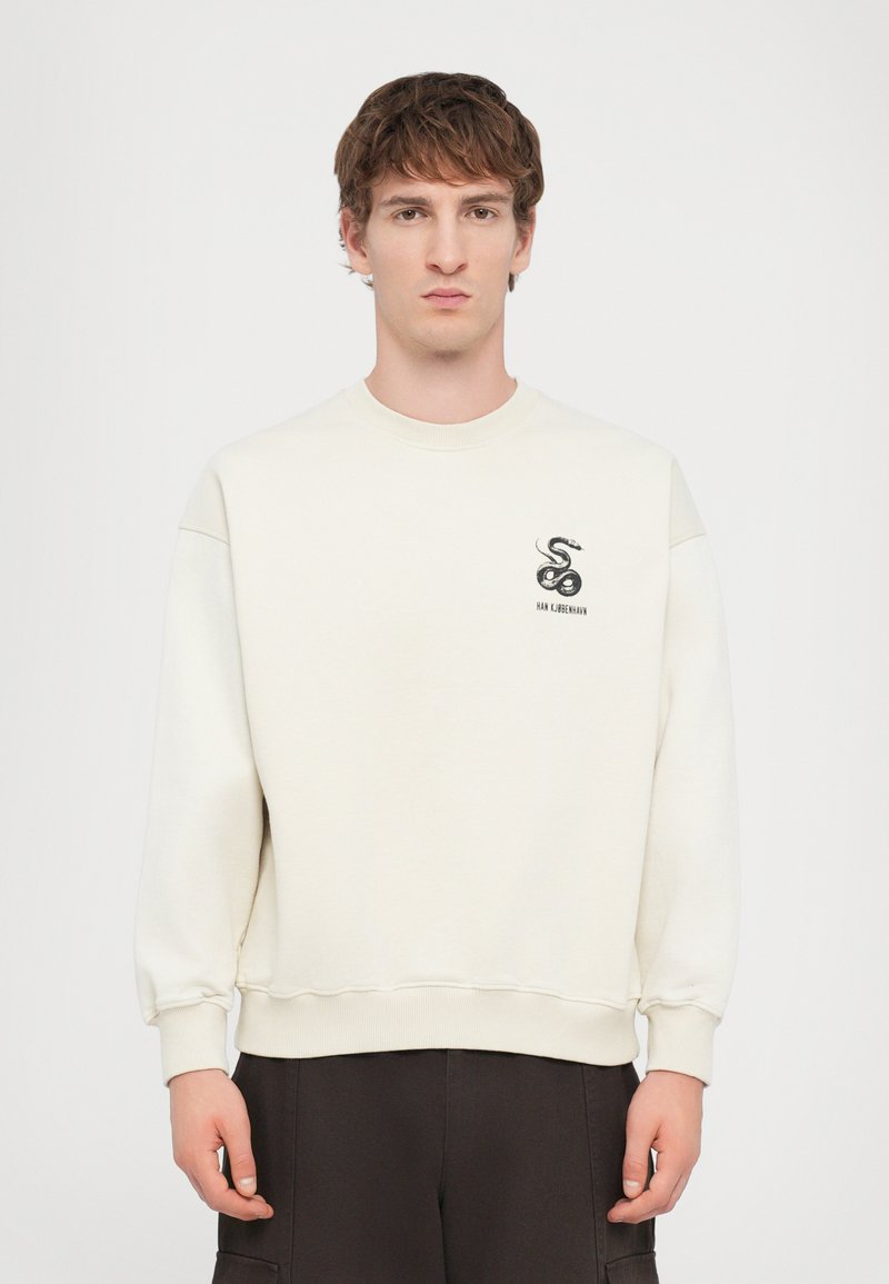 Cremefarvet sweatshirt lavet af blødt materiale, med et lille sort slange-logo og ribbede manchetter og kant, båret med mørke bukser.