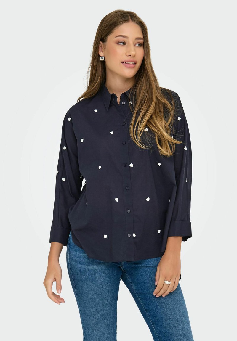 Navy button-up shirt with white heart patterns, long sleeves with rolled cuffs, and a relaxed fit. Paired with blue denim jeans.