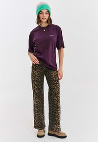 Žena oblečená v purpurovém tričku s malým designem řasy, leopardím potiskem na kalhotách, béžovými botami a růžovo-zelenou čepicí, stojící na bílém pozadí.