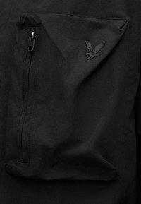 Μαύρη τσέπη από ύφασμα με κάθετο φερμουάρ στα αριστερά και ανάγλυφο λογότυπο αετού στην ανυψωμένη δεξιά πλευρά της τσέπης.