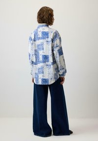 Cămașă albă cu modele paisley și geometrice albastre; croi lejer cu mâneci lungi. Asortată cu pantaloni din denim închis cu croială largă.