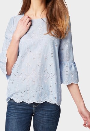 Femme portant un chemisier en dentelle ajourée bleu clair avec un ourlet festonné et des manches cloches trois-quarts, associé à un jean bleu foncé.