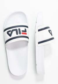 Vita glid sandaler med en texturerad sula, ett brett band med svart och röd FILA-logotyp och en övergripande slät, minimalistisk design.