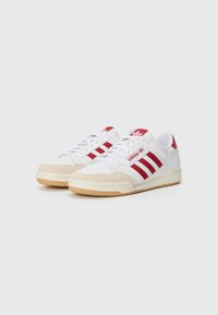 Zapatillas blancas con rayas rojas, materiales de ante y cuero, punta redonda, cordones blancos, suela de goma y logotipo de marca prominente en la lengüeta y el talón.