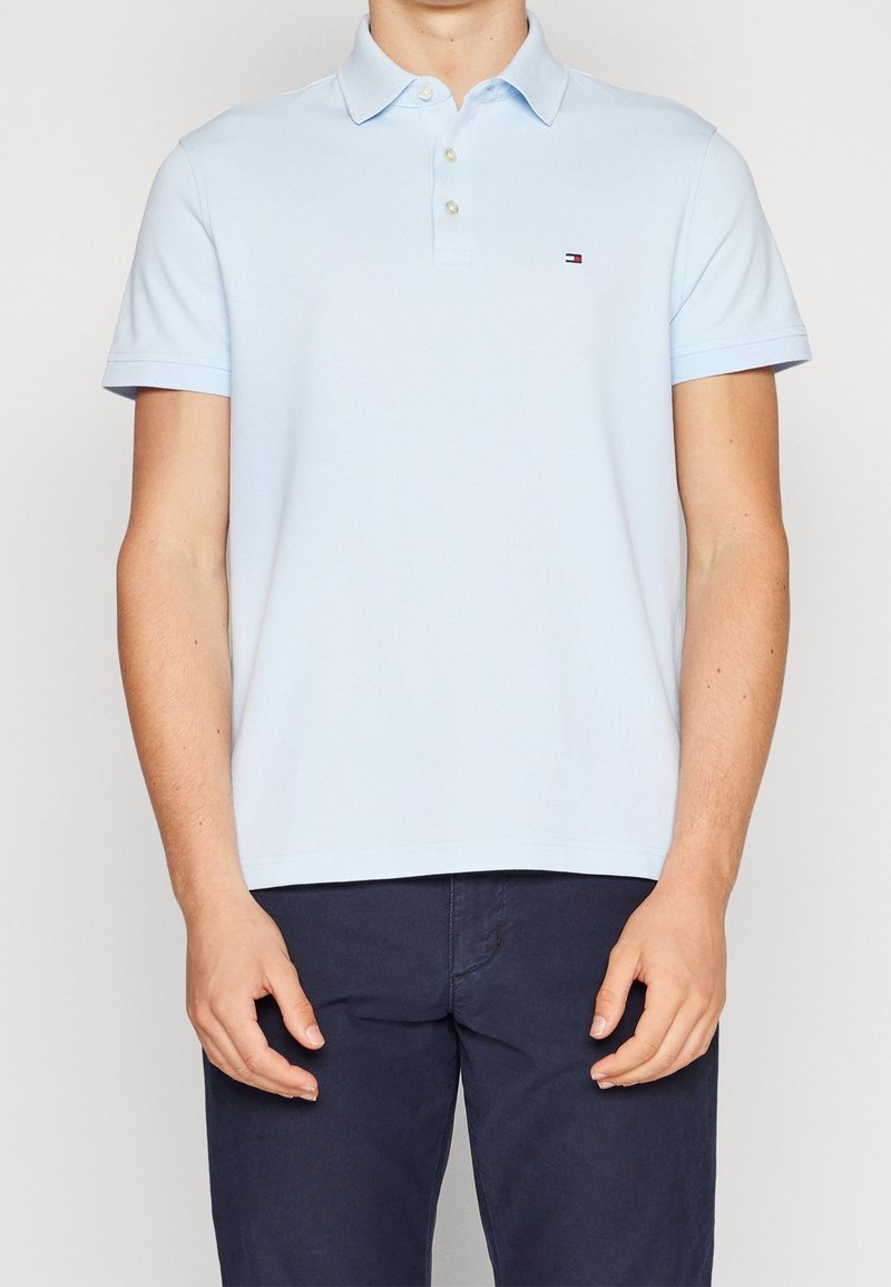 Homme portant un polo bleu clair à manches courtes et un pantalon bleu foncé, debout, les bras détendus le long du corps, sur un fond uni.