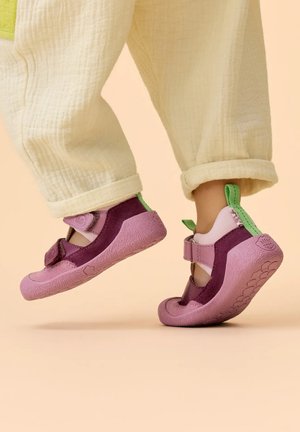 Voeten van een kind, gedragen in paarse en roze schoenen met zachte zolen en groene lusjes, staand op de tenen tegen een beige achtergrond.