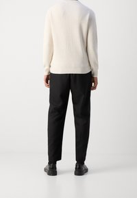 Off-white gerippter Strickpullover kombiniert mit schwarzen Hosen und schwarzen Schuhen, von hinten gesehen, mit Betonung auf Textur und Farbkontrast.