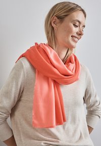 Sciarpa corallo realizzata in morbido materiale, adagiate attorno al collo con bordi frangiati, abbinata a un maglione beige chiaro.