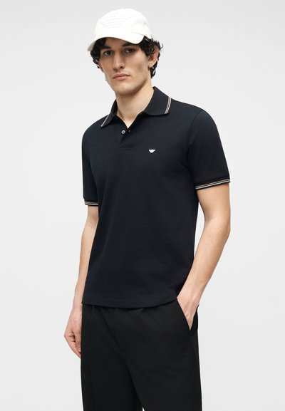 Νεαρός άνδρας που φοράει μαύρο πόλο με ριγέ γιακά και μανίκια, μαύρο παντελόνι, λευκό καπέλο, στέκεται με το ένα χέρι στην τσέπη.