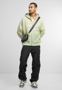Sweat à fermeture éclair vert avec le texte "Retinality", sac bandoulière noir, pantalon cargo noir et baskets multicolores, sur un fond neutre.