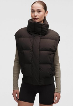 Mujer con el pelo recogido, vistiendo una chaqueta negra acolchada sin mangas sobre una camiseta beige de manga larga y pantalones cortos negros, de pie frente a un fondo gris.