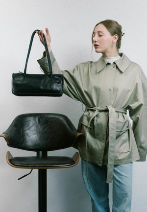 Zwarte leren handtas met een rechthoekige vorm en twee lange hengsels. Model draagt een lichtgrijze, ceintuurloze jas over een blauwe spijkerbroek, naast een donkere leren stoel.