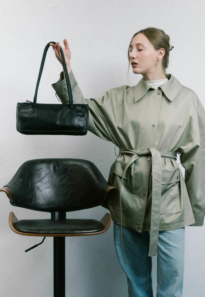 Sac à main en cuir noir de forme rectangulaire avec deux longues poignées. Le modèle porte une veste gris clair ceinturée sur un jean bleu, à côté d'un fauteuil en cuir foncé.