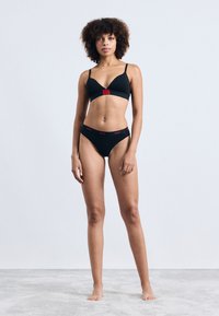 Set de sutien negru și bikini asortat, cu un bandou cu logo roșu. Sutienul are bretele subțiri și o formă triunghiulară. Textura fabricii este netedă.